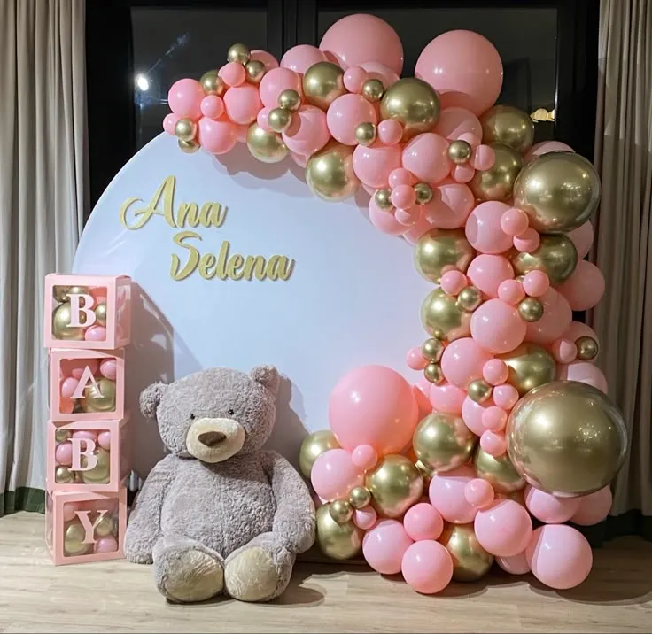 Gender reveal baby setup with round backdrop, golden and baby pink balloons, baby name box, and a medium-size teddy bear.